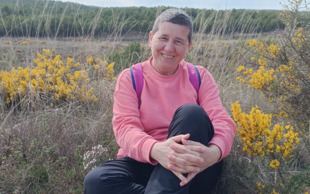 Anaís Isarre sentada sonriente en un campo de flores silvestres celebrando el inicio de la primavera y el Día de la Felicidad en su ruta por el río Aguasvivas.